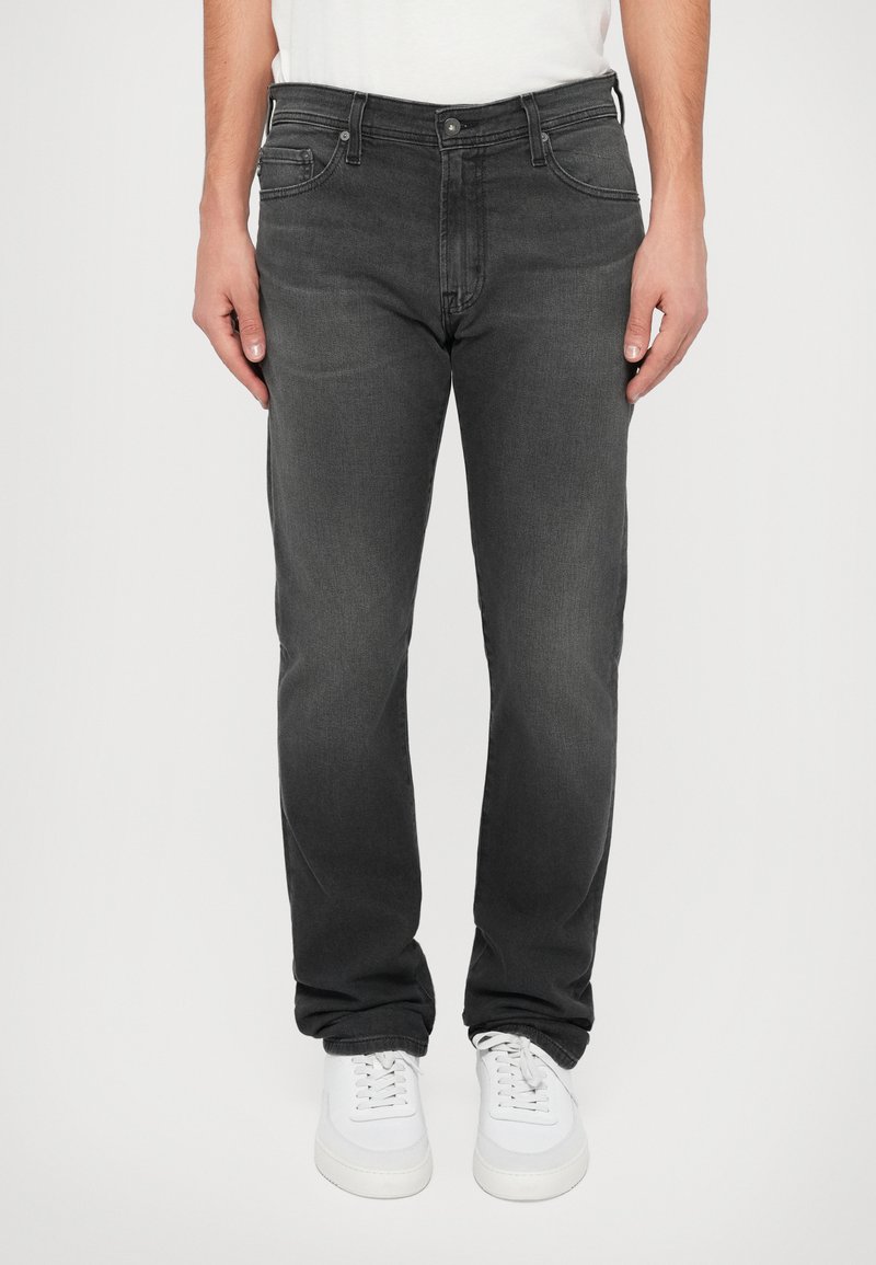 Jeans de ganga cinza escuro com corte reto, cinco bolsos e um acabamento ligeiramente desbotado, combinados com ténis brancos.