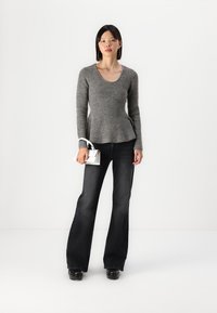 Grå strikket genser med rå neckline, kombinert med svarte flared jeans. Modellen holder en liten hvit veske, og står mot en ensfarget bakgrunn.