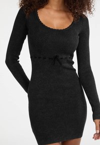 Femme portant une robe moulante noire en maille côtelée à manches longues avec un petit nœud sous la poitrine.