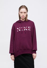 Vijolična kapuca z žepom spredaj, ki vsebuje bel Nike logotip in rdeč napis. Oblikovanje ima ohlapen kroj in kapuco z vrvico.