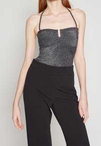 Kobieta w błyszczącej srebrnej bluzce na halter z centralnym wycięciem i czarnych spodni high-waisted na jednolitym tle.