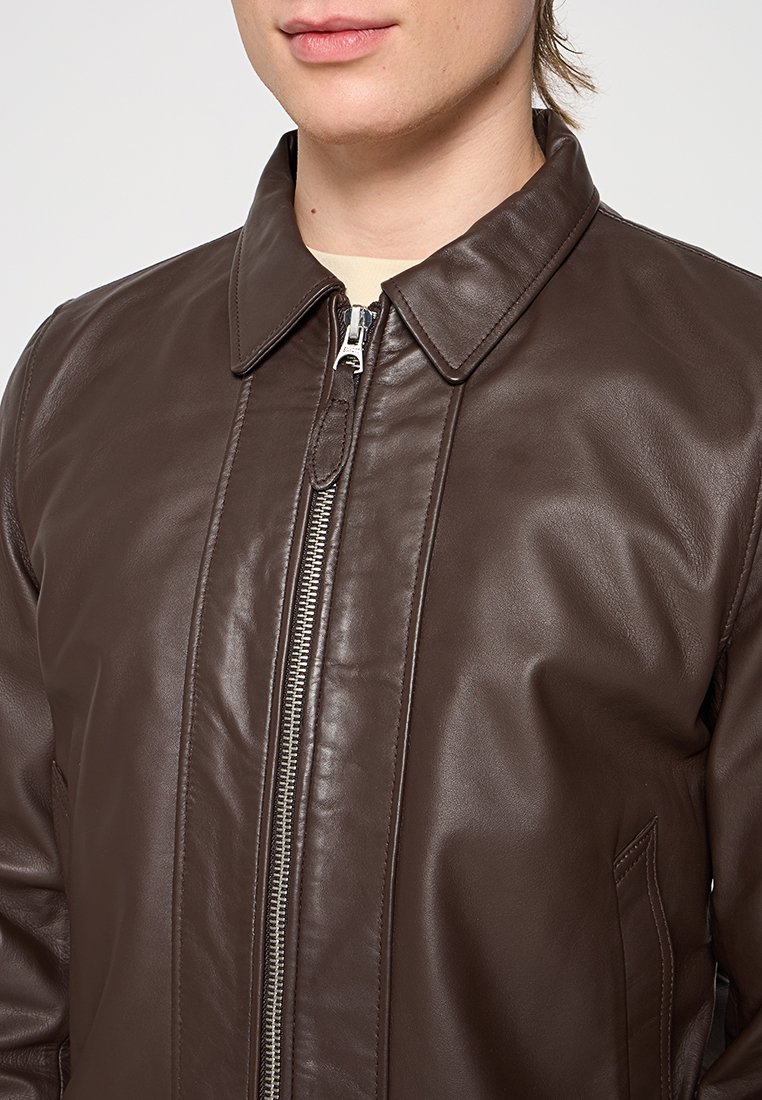 Veste en cuir marron avec une fermeture éclair frontale, un col pointu et des poches latérales. La texture est lisse, avec des détails de couture subtils.