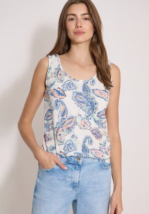 Femme portant un haut sans manches à motif cachemire blanc, bleu et pêche, associé à un jean bleu clair, debout avec une main dans la poche.