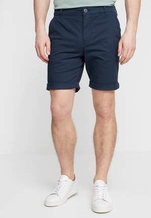 Marineblaue Baumwollshorts mit umgeschlagenen Säumen, ausgestattet mit einem vorderen Knopfverschluss und Seitentaschen, kombiniert mit weißen Turnschuhen.