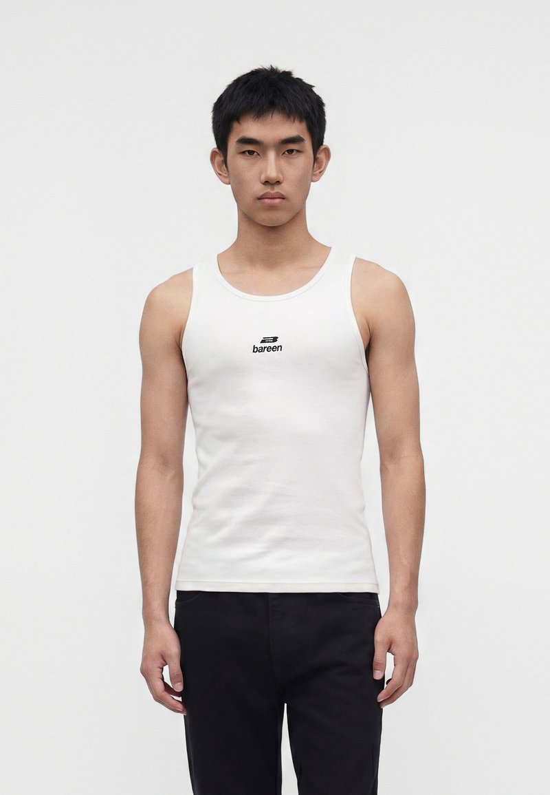 Jeune homme debout, face à l'objectif, portant un débardeur blanc avec le logo "bareen" et un pantalon noir, sur un fond blanc uni.