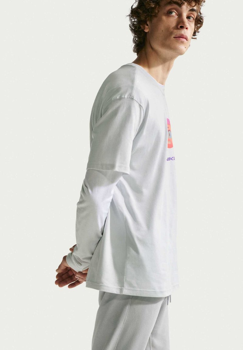 Joven con pelo rizado que lleva superpuestas camisas blancas de manga larga y corta con gráfico, de pie con las manos detrás de la espalda.