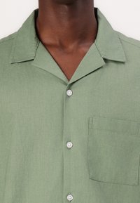 Camisa verde de manga corta y botón, hecha de una tela ligera, con un único bolsillo en el pecho y botones plateados. Textura suave.