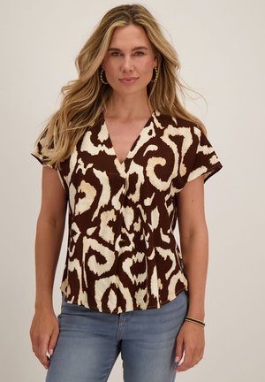 Vrouw met lang blond haar, gouden oorringen, een bruin-beige blouse met korte mouwen en patroon, en een blauwe spijkerbroek, staand tegen een effen achtergrond.