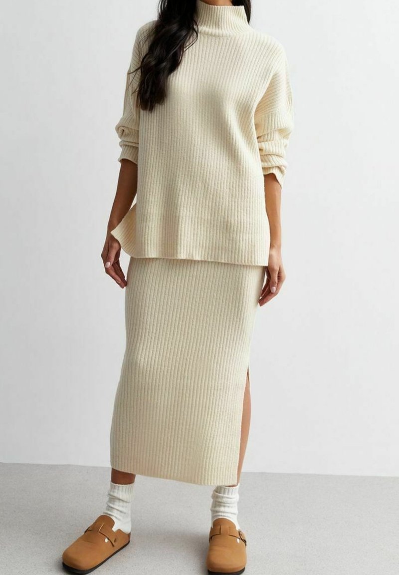 Femme portant un pull à col roulé en maille côtelée crème et une jupe midi assortie avec une fente latérale, associées à des chaussettes blanches et des chaussures plates beiges.