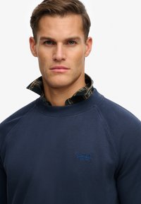 Felpa girocollo blu navy con tessuto strutturato e logo blu. Indossata sopra con un colletto di camicia a quadri verde e marrone che sporge sotto.