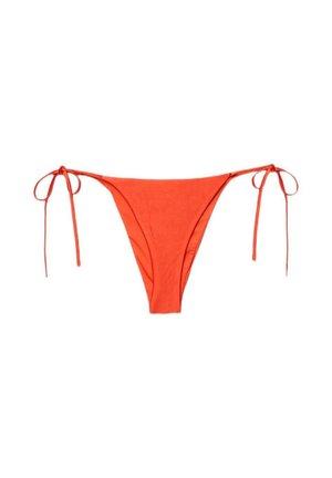 Donna con i capelli bagnati che aggiusta la parte inferiore del bikini rosso con laccetti su uno sfondo marrone uniforme.