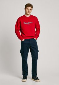Sudadera roja con logo, pantalones cargo azul marino y zapatillas negras con suelas beige. El modelo está de pie con las manos en los bolsillos sobre un fondo neutro.