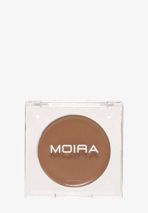Pieni pyöreä meikkituote keskiruskeassa sävyssä, läpinäkyvässä neliönmuotoisessa kotelossa, jossa valkoinen "MOIRA"-logo päällä.
