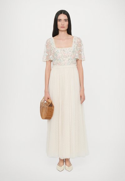 Maxi-jurk met een doorzichtig lijfje met bloemenborduursel, een geplooide rok in ivoorkleur en een geweven handtas van bamboe. Puntige witte schoenen maken de look compleet.