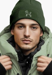 Green knit beanie with a textured finish and a black patch featuring a dog graphic, worn over a padded green jacket.