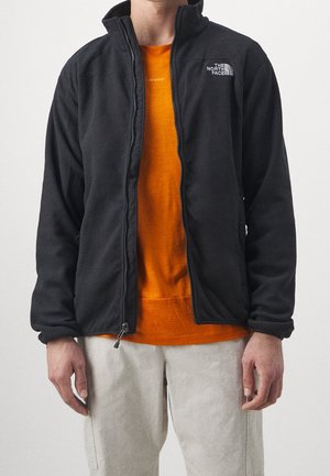 Homme portant une veste polaire noire ouverte sur une chemise orange avec un pantalon clair sur fond uni.