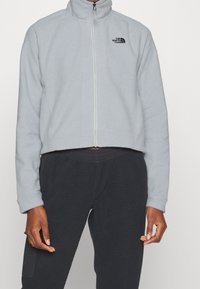 Jasnoszara polarowa kurtka z wysokim kołnierzem, pełnym zamkiem błyskawicznym i logo The North Face na lewej piersi. Krótkie cięcie, w połączeniu z czarnymi spodniami.