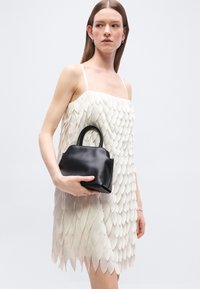 Femme en robe blanche texturée sans manches tenant un petit sac à main en cuir noir avec poignées supérieures sur un fond uni.