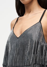 Μαύρο τοπ με V-λαιμόκοψη και λεπτές τιράντες, διακοσμημένο με μεταλλικές παγιέτες και φουντίτσες, δημιουργώντας μια υφή με ανακλαστική επιφάνεια.