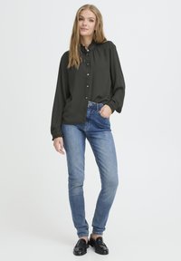 Tumesinine bluus kõrge kaelusega ja nööpidega, koos helesiniste kitsaste teksadega. Mudel kannab musti mokasid; keskkond on neutraalne.