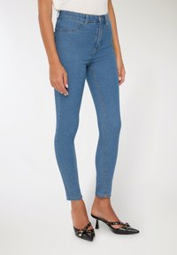 Kobieta w obcisłych niebieskich dżinsach i czarnych czółenkach na obcasie z ostro zakończonym noskiem, ozdobionych złotymi ćwiekami, na białym tle.