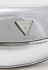 Bolsa prateada em imitação de couro com um log�ótipo metálico triangular contendo o texto "GUESS" e um ponto de interrogação. Textura suave com costuras decorativas.