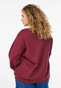 Sweatshirt bordeaux avec une coupe décontractée, un col rond et des manches longues. Tissu doux avec des coutures visibles et des poignets côtelés au bas.