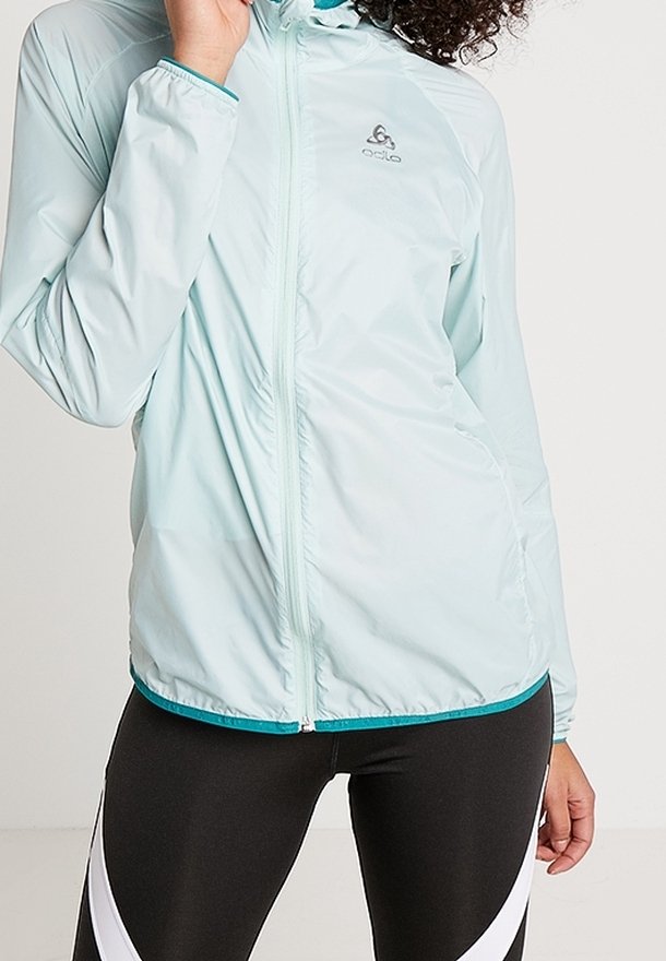 Femme portant une veste zippée bleu clair et des leggings noirs avec des rayures blanches diagonales, main près du col.