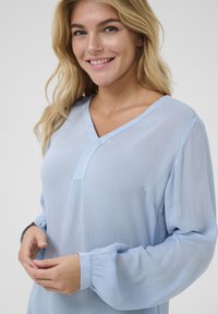 Blouse bleu clair en coupe décontractée, avec un col en V, de longues manches bouffantes et un tissu texturé avec des poignets froncés.