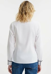Camisa blanca de manga larga con corte recto, con ribetes negros en los puños y una textura de tela suave, combinada con jeans azules.