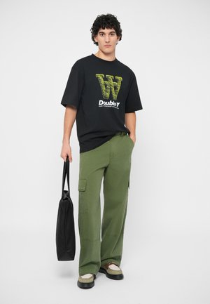 Μαύρο oversized t-shirt με κίτρινο γραφικό "W" και κείμενο, συνδυασμένο με πράσινα cargo παντελόνια. Το μοντέλο κρατάει μια μαύρη τσάντα tote, φορώντας μπεζ σανδάλια.