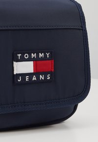 Borsa in poliestere blu navy con patta. Presenta un'etichetta rettangolare con il testo "TOMMY JEANS" e un design a blocchi rosso, bianco e blu. Finitura in tessuto testurizzato.
