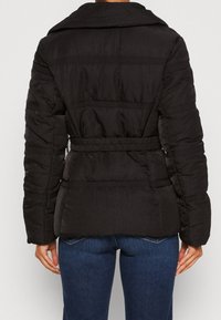 Chaqueta negra acolchada con un cinturón en la cintura, llevada por una persona con jeans azules, vista desde la espalda.