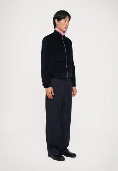 Veste en velours côtelé marine avec poches avant, fermeture éclair argentée, portée sur un col roulé rose. Associée à un pantalon large à rayures marines et des chaussures noires.
