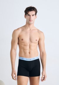 Man with short curly hair wearing black Tommy Hilfiger boxer briefs with light blue waistband standing against plain white background.