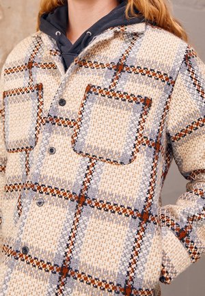 Geruite shacket in beige, grijs en oranje. Bevat twee grote zakken en zwarte knopen, gemaakt van geweven textuurstof.