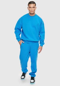 Blå hettegenser og matchende joggebukser laget av bomullsblanding, med en avslappet passform, ribbet mansjetter, og svarte og hvite sneakers.