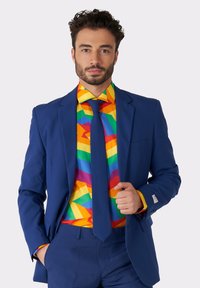 Blauwe blazer met een bijpassende stropdas en een kleurrijk geometrisch patroon op het shirt met felle kleuren zoals geel, rood en groen.