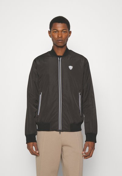 Veste bomber noire avec fermeture éclair blanche et accents, fabriquée en tissu lisse. Présente un logo sur la poitrine et des poignets et un ourlet côtelés.