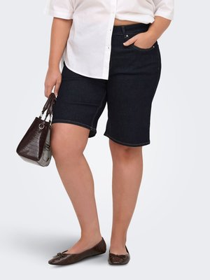 Persoon draagt donkere denim knielange shorts, wit overhemd met knopen, bruine platte schoenen, houdt een bruine handtas vast met één hand in de zak.