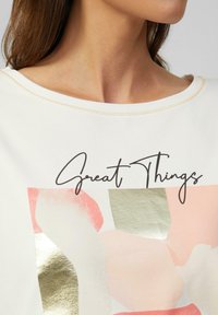 Witte top met een ronde halslijn, voorzien van een kleurrijke print van abstracte vormen in roze, perzik en groen, met een gouden accent.
