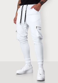 Weiße Jogginghosen aus einem weichen Stoff mit elastischem Bund und schwarzen Kordeln. Ausgestattet mit seitlichen Reißverschlusstaschen und gerippten Bündchen. Kombiniert mit weißen Sneakers.