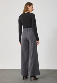 Black long-sleeve top paired with high-waisted, wide-leg gray trousers. Smooth fabric with an elastic waistband and subtle sheen. Black high heels.