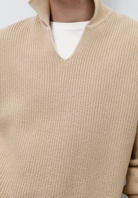 Beżowy sweter dzianinowy z dekoltem w kształcie litery V i półzamkiem. Tekstura jest prążkowana, a pod spodem widoczna jest warstwowa biała koszulka.