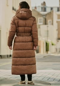 Abrigo puffer acolchado largo y marrón con capucha, cinturilla elástica y abundante relleno, que se lleva sobre pantalones negros y zapatillas de color claro.
