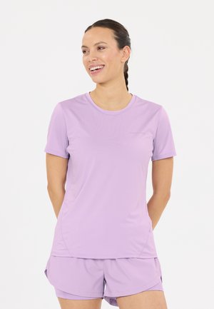 Frau mit dunklem geflochtenem Haar lächelt, trägt ein hellviolettes kurzärmeliges Shirt und passende Shorts, steht vor einem schlichten weißen Hintergrund.
