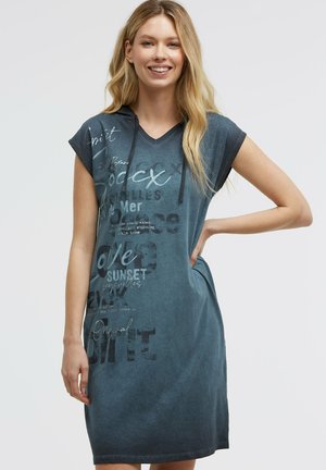 Lächelnde Frau mit langen blonden Haaren, die ein blaues kurzärmeliges Kapuzenkleid mit bedrucktem Text trägt, steht mit einer Hand in der Hüfte.