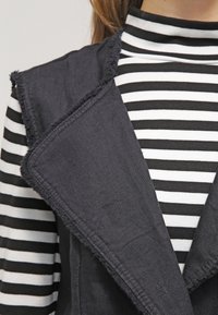 Un gilet noir avec un col effiloché, superposé sur un col roulé à rayures noires et blanches. Les deux tissus semblent lisses, et le gilet a un design ouvert.