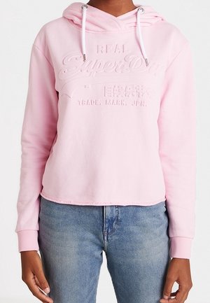 Hellrosa Hoodie mit geprägtem "SuperDry"-Logo und japanischem Text, kombiniert mit blauen Denim-Jeans.