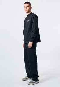Camisa de algodón negra de manga larga con cuello redondo, con texto blanco en el pecho, combinada con pantalones negros holgados y zapatillas grises.